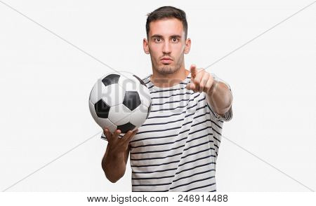Handsome young man holding soccer football pointing with finger to the camera and to you, hand sign, positive and confident gesture from the front