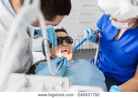 medicine, dentistry and healthcare concept - dentist and assistant with dental drill and saliva ejector treating kid patient teeth at dental clinic