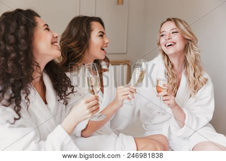 Photo of joyful three women 20s wearing housecoat smiling and lying in big bed in luxury apartment or hotel room with glasses of champagne during hen party