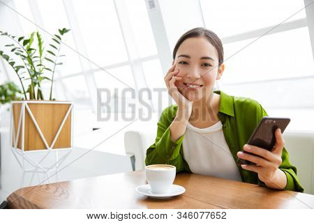 Smiling asian woman using mobile phone while sittng at the cafe indoors and drinking coffee