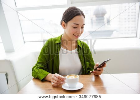 Smiling asian woman using mobile phone while sittng at the cafe indoors and drinking coffee