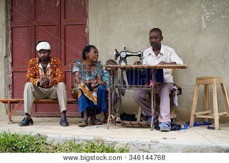 December, 2019. Africa, Uganda, A Man Sews On An Old Mechanical Sewing Machine And Two People Waitin