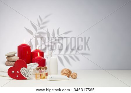 Spa still life with creams, essential oils, burning red candles, hearts on light background with shadow from plant. Healthy lifestyle, body care, Spa treatment. Valentine's Day. Honeymoon. Wedding