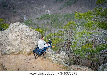 Travel Man Working With Laptop Sitting On The Rocky Mountain On Beautiful Scenic Clif Background