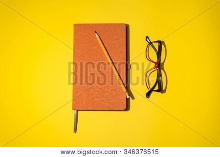 A Pencil, Orange Book And Red Black Glasses On The Yellow Background.