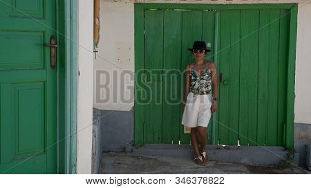 Asian Woman In Sunglasses And Hat Standing In Front Of Traditional Greek House With Green Shutters A