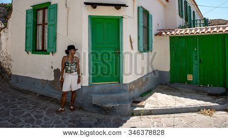 Asian Woman In Sunglasses And Hat Standing In Front Of Traditional Greek House With Green Shutters A
