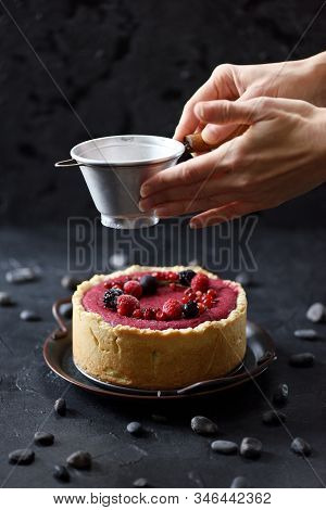 Decorating Homemade Pie. Woman Hands Pouring Icing Sugar On Open Red Berry Pie On Black Background H