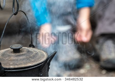 Close-up Of A Sooty Teapot With A Lid Hanging And Basking Over A Campfire, On A Blurry Background.