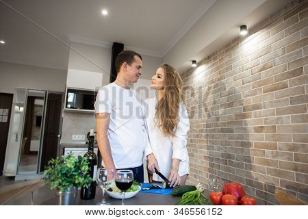 Portrait Of Omantic Couple In The Kitchen At Home. Blonde Woman With Long Hair Slices Cucumber. A Ma