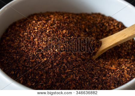 Close Up Shot Of Dried Rooibos (redbush) Tea Leaves In A White Ceramic Bowl With Wooden Spoon.
