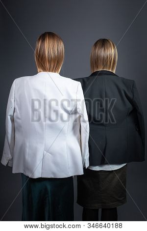 Two Girls With Blond Hair Tuckin Jackets Standing Against Gray Background In Strict Suits, Rear View