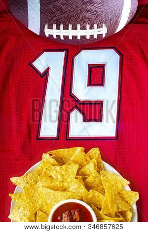 An American Football With Organic Nacho Chips And Mild Salsa On A White Red Football Jersey With The