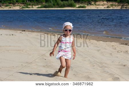 Capricious And Unhappy Caucasian Child Of Three Years In Sunglasses Walking On Sand River Beach