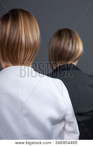 Two Girls With Blond Hair Tuckin Jackets Standing Against Gray Background In Strict Suits, Rear View