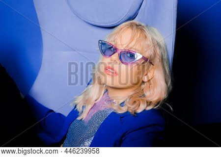 A Girl In A Blue Jacket And Purple Glasses Sits On A Chair. Fashion Photo In 80s 90s Style.