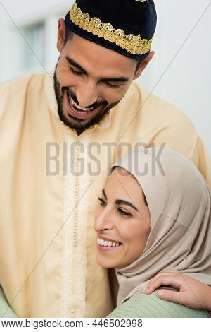 Cheerful Mislim Man In Skullcap Laughing Near Young Happy Wife At Home