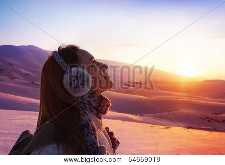 Image of pretty woman walking in snowy mountains, side view of cute girl looking up, closeup portrait of female wearing warm winter earmuff, red sunset, wintertime sports, trekking and hiking concept