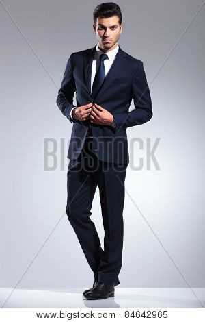 Full body picture of a young business man closing his jacket while looking at the camera.