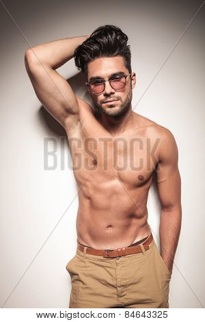 Hot sexy young man leaning on a white wall while holdinh one hand on his heair and the other one in his pocket.