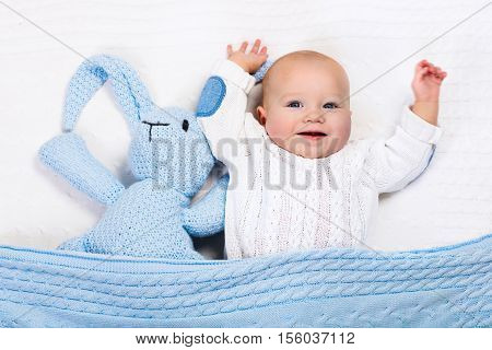 Funny little baby wearing a warm knitted jacket playing with toy bunny relaxing on white cable knit blanket in sunny nursery. Kids winter clothing and bedding. Hand made toys and textile for children.
