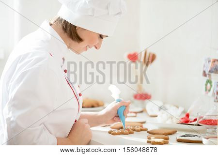 Professional smiling female confectioner decorating a gingerman with icing sugar using a pastry bag, preparing for Christmas. Lifestyle, horizontal, Christmas concept photo