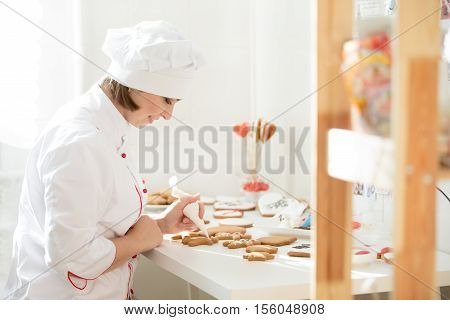 Professional female confectioner sitting and decorating a gingerman with icing sugar using a pastry bag, preparing for Christmas. Lifestyle, Christmas concept photo