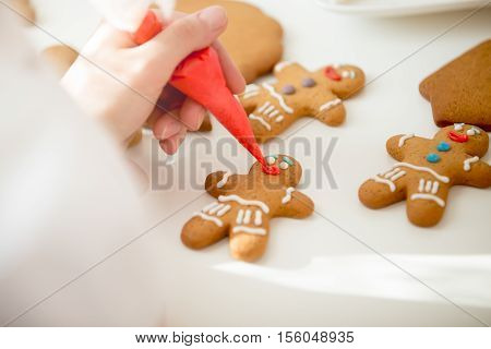 Close up of confectioner hand decorating a gingerman with a pastry bag, drawing a smile, making it cute, fun and delicious View over a shoulder, lifestyle
