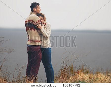 Young attractive couple in knitted sweaters standing on a cliff at sea shore and hugging