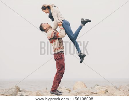 Young attractive couple in knitted sweaters on a cliff on sea shore man hold his girfriend on his hands in the air creative toning