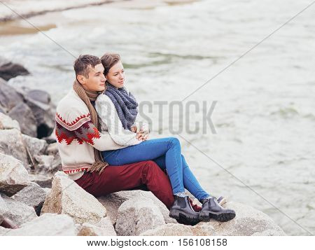 Young attractive couple in knitted sweaters on a cliff on sea shore hugging and holding hands creative toning