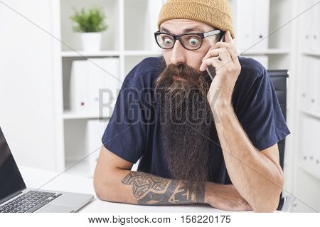 Baffled long bearded hipster is talking on the phone at his office. The laptop is standing on the desk
