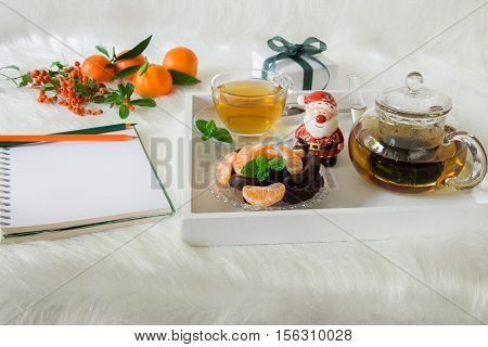 Green mint tea, chocolate, mandarin slices and Santa Claus on tray near notebook, pencils, mandarins, mountain ash on white artificial fur background. Time break notebook surprise preparation.