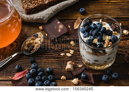 Granola with yogurt blueberries honey and chocolate bar on wooden table with ingredients.