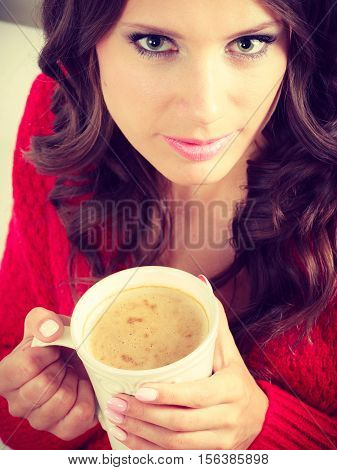 Attractive fall girl red autumnal sweater holding white mug with coffee warm beverage sitting on sofa at home. Woman warming herself relaxing top view