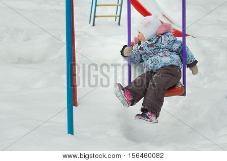 Little girl is riding on frosty winter playgrounds swing is covering with white snow outdoors
