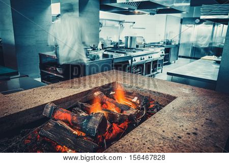 Restaurant kitchen interior: brazier with burning wood, made of natural stone with fire for BBQ. In the background buzzing restaurant work motion chefs