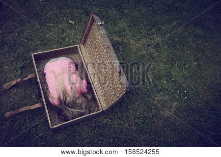 Child blond girl inside a suitcase on green grass lawn. Concept image of refugee orphan or poor child.