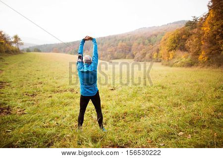Senior runner in sunny autumn nature doing stretching. Man with smart phone in armband on his arm. Rear view.