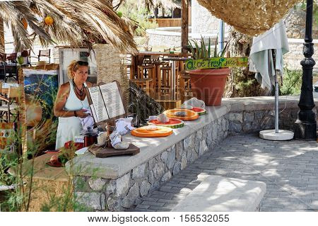 PALEOCHORA, CRETE, GREECE - JULY 2016: Woman offering products at street cafe in Paleochora town on Crete island