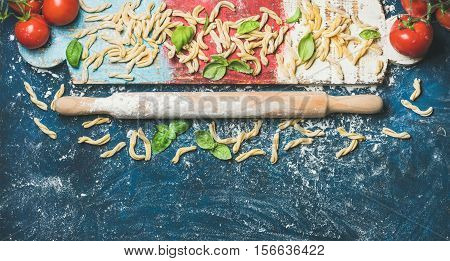 Ingredients for cooking Italian dinner. Fresh pasta casarecce, cherry-tomatoes, basil leaves and plunger on colorful wooden board over dark blue background. Top view, copy space. Food frame concept