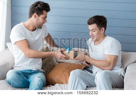 Non-traditional relationship. Handsome relaxed joyful man smiling and accepting a gift from his male friend while sitting together on a sofa at home.