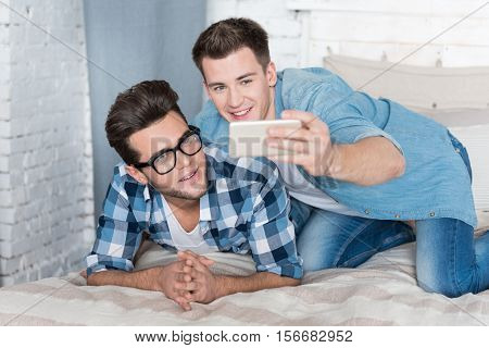 Having fun together. Joyful handsome gay couple smiling and making selfies together while relaxing on a bed.