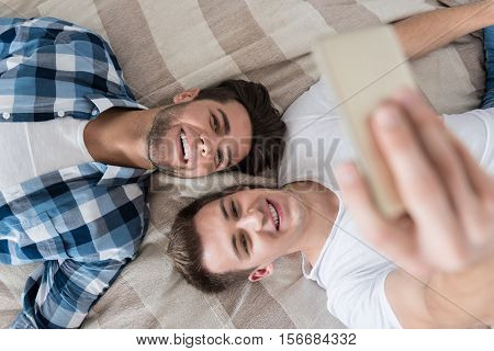 Best memories. Delighted young gay couple making selfie with smartphone while lying in bed together and smiling.