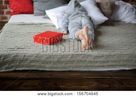 The child in pajamas lies on the big bed with his face buried in the pillow. Nearby there is a bright red box with a gift. The child isn't happy. He is disappointed with a gift and doesn't want to look at this gift. Offense. Protest.