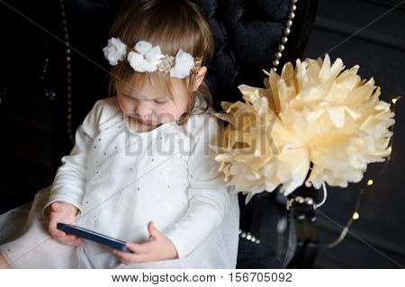 Bonny ruddy toddler sitting in the chair and enthusiastically looking at smartphone screen. In white dress with a wreath on his head the child like an angel.