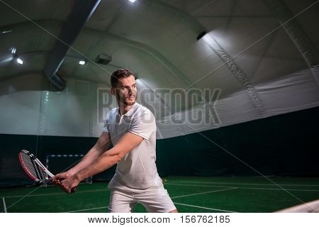 Train your skills. Pleasant profesional concentrated man holding racket and playing tennis while training in indoor tennis court