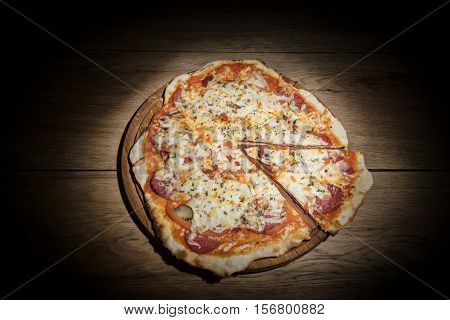 Delicious round pizza with salami and pickles and with slice cut out served on a wooden plate on old wood table in the dark