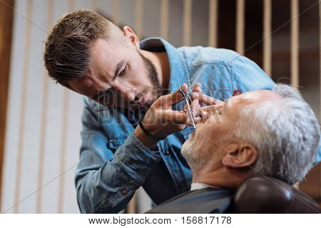 Very scrupulous. Attractive bearded hairstylist cutting mustache of senior client sitting at barbershop.