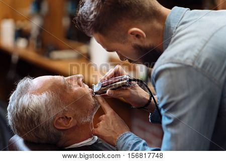 Precise work. Side view of young attractive bearded hairstylist trimming beard of old barbershop visitor.